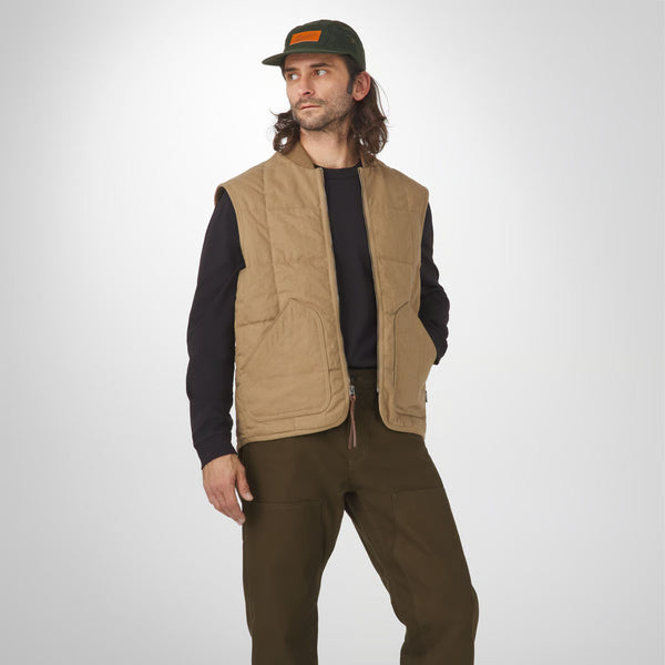 Man wearing a tan vest over a black shirt and brown pants on a white background