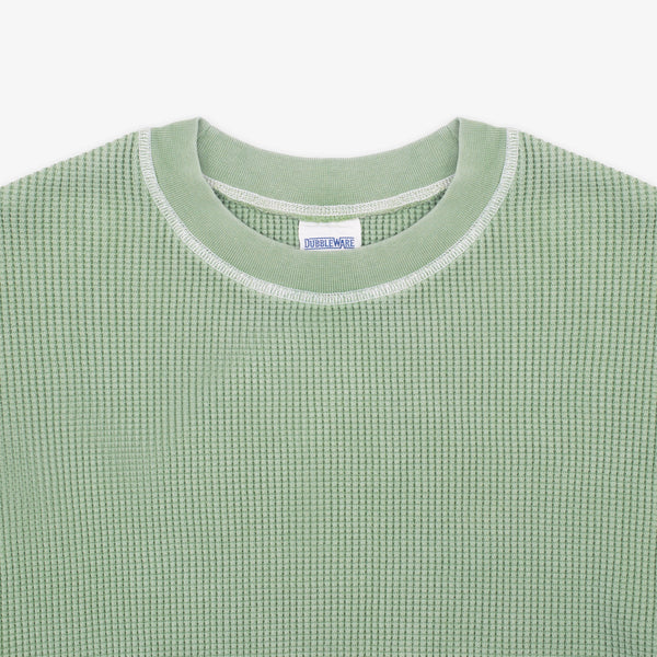 Close-up of a light sage green waffle-knit crewneck sweater with a white ribbed collar trim and a visible neck label reading 'SWEATWEAR'.