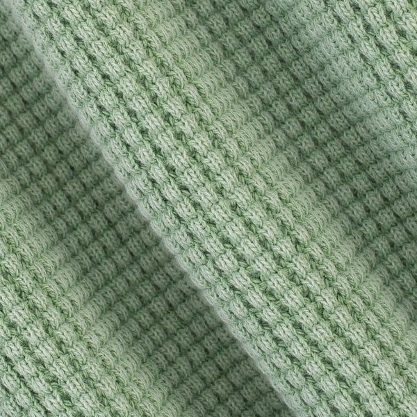 A close-up of a light sage green knit fabric with a textured, ribbed pattern and soft diagonal folds.