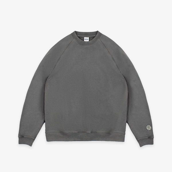 A dark gray crewneck sweatshirt with raglan sleeves, laid flat against a light gray background. A small circular logo patch is visible on the left sleeve cuff.