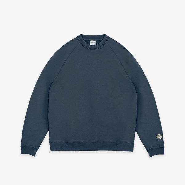 A dark slate blue crewneck sweatshirt with raglan sleeves, shown flat against a light gray background. A small circular logo is printed on the left sleeve cuff.