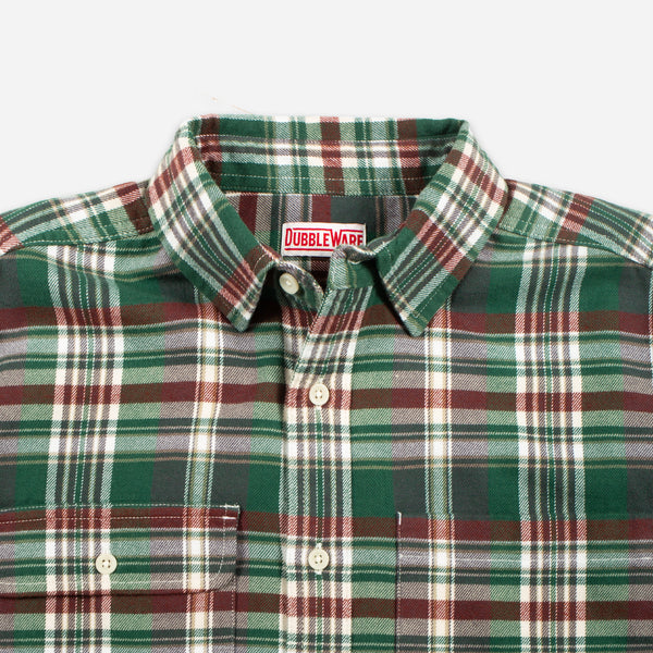 A close-up of a Dubbleware plaid button-down shirt in green, burgundy, and white, showing the collar, buttons, and chest pocket.