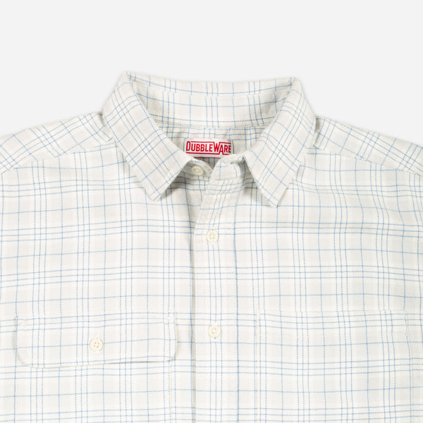 Close-up of a Dubbleware button-down shirt with a white base, light blue windowpane plaid, and cream buttons, featuring a collar and chest pocket.