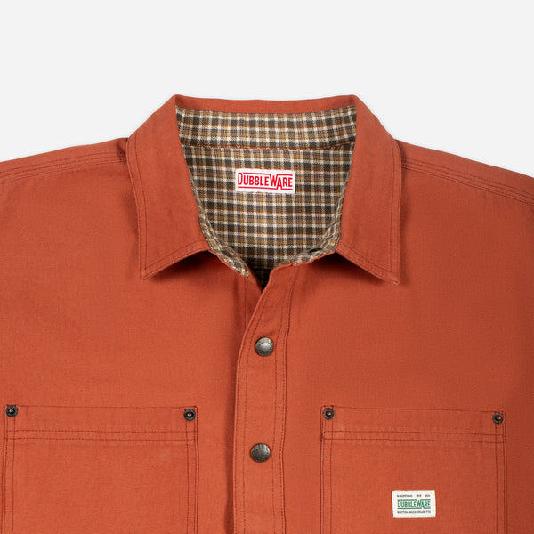 Close-up of a Doubleware work shirt in rust orange, showing the collar with a brown plaid lining, snap-button front, and a small white brand patch on the chest pocket.