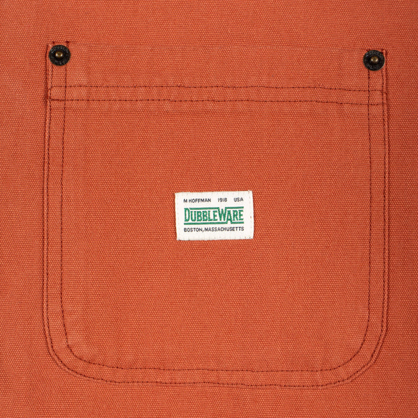Close-up of a textured orange canvas pocket with a white Dubbleware label stitched in the center, featuring two black snap buttons at the top corners.