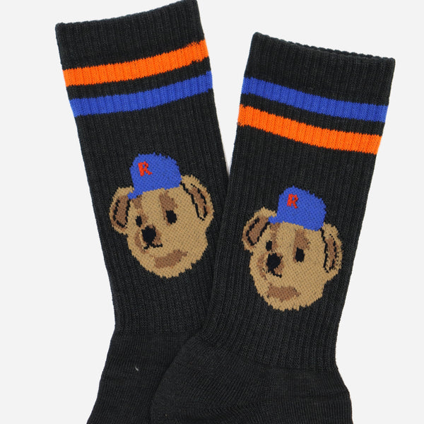 A pair of black crew socks featuring a knitted illustration of a puppy wearing a blue cap with a red letter 'B' on the side, and two horizontal stripes near the cuff in blue and orange.