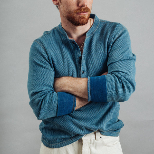 A man with red hair and a beard wearing a textured blue henley shirt with contrasting dark blue cuffs, posed with arms crossed against a solid gray background.