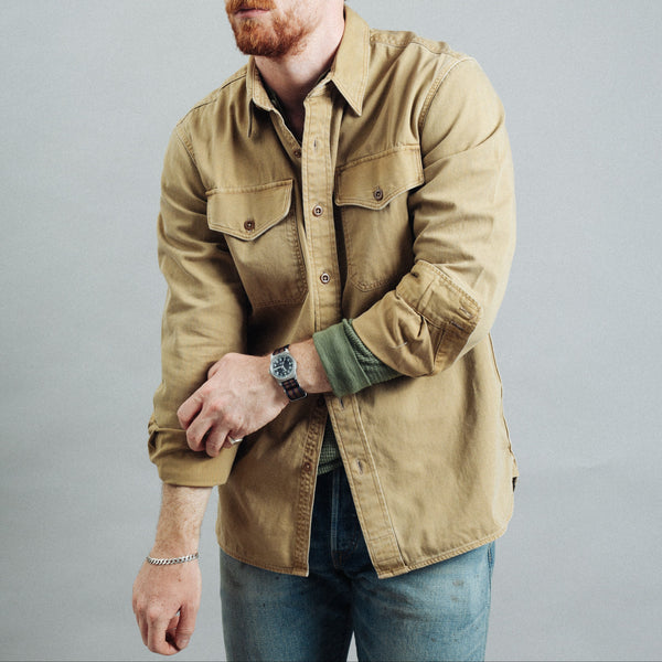 A man wearing a tan utility button-up shirt with a green undershirt, blue jeans, and a brown cap, posed against a solid gray studio background.