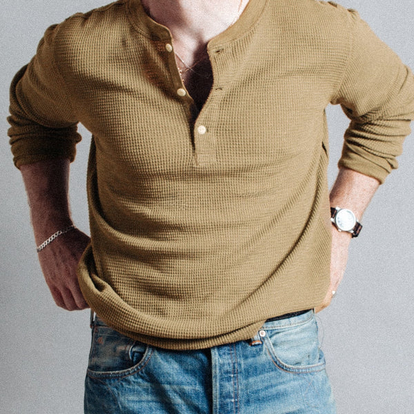 A man wearing a textured, long-sleeve Henley shirt in a warm olive-brown color, tucked into blue jeans, shown from the chest down against a neutral gray background.