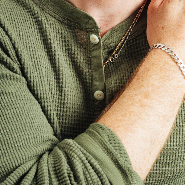 Close-up of a person wearing an olive green waffle-knit long-sleeve henley shirt, with the collar open, buttons visible, and one arm raised wearing a silver chain bracelet.