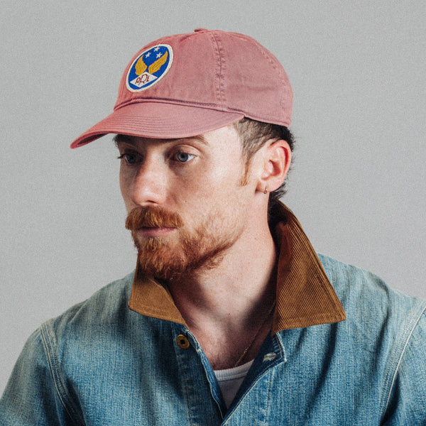 A man with a red beard wearing a dusty rose baseball cap with a circular blue, yellow, and white logo patch, paired with a blue denim shirt jacket with a brown corduroy collar, set against a plain gray background.