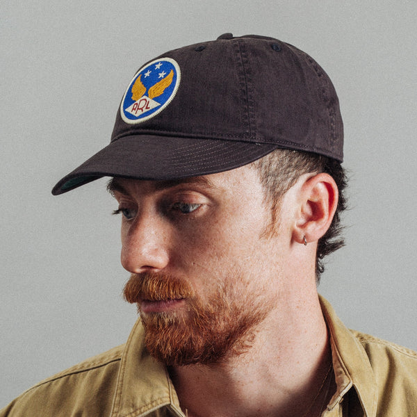 A man wearing a dark brown baseball cap with a circular embroidered patch featuring an eagle and stars, paired with a tan button-up shirt against a plain gray background.