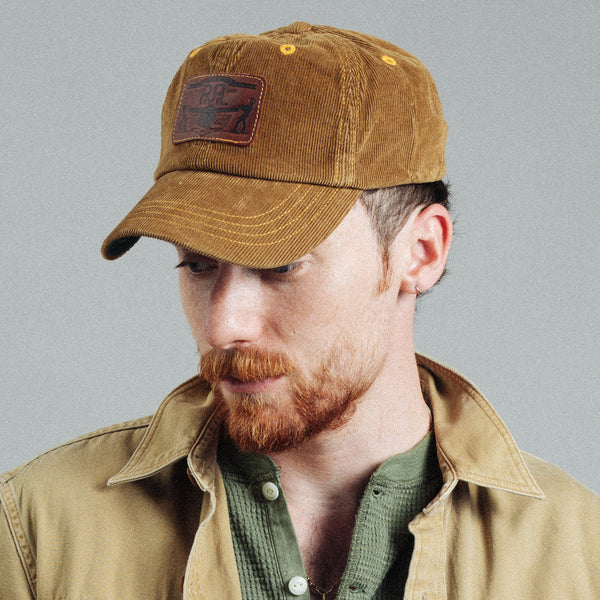 A man wearing a brown corduroy baseball cap with a brown leather patch that reads 'SUNRISE', paired with a tan overshirt and green shirt, against a plain gray background.